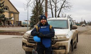 A World Food Programme worker stands in front of a muddy white SUV, holding a blue helmet, in a Ukrainian town.