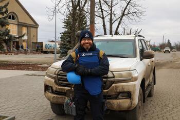 A World Food Programme worker stands in front of a muddy white SUV, holding a blue helmet, in a Ukrainian town.