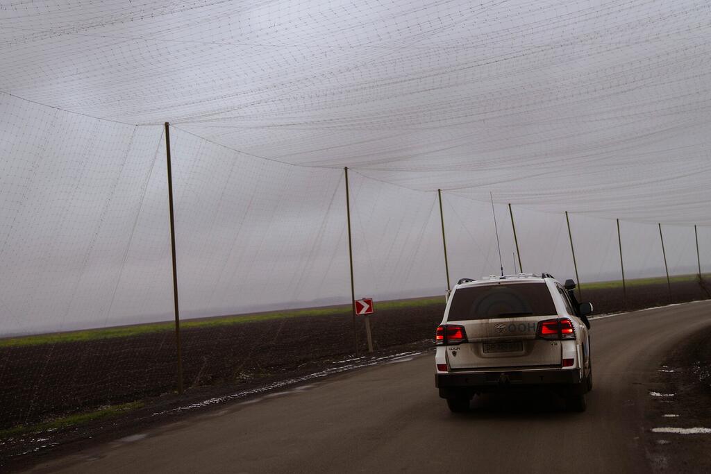 Un SUV blanco conduce a lo largo de una carretera rural en Ucrania, pasando bajo una gran red agrícola apoyada por postes.