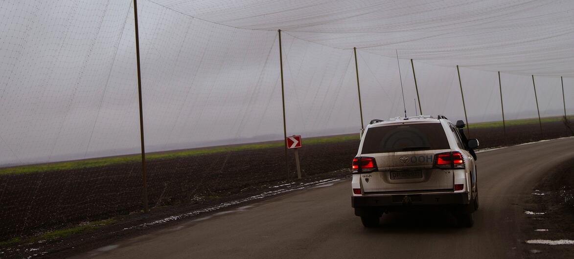 A white SUV drives along a rural road in Ukraine, passing under a large agricultural net supported by poles. The scene suggests humanitarian or emergency response operations by WFP.