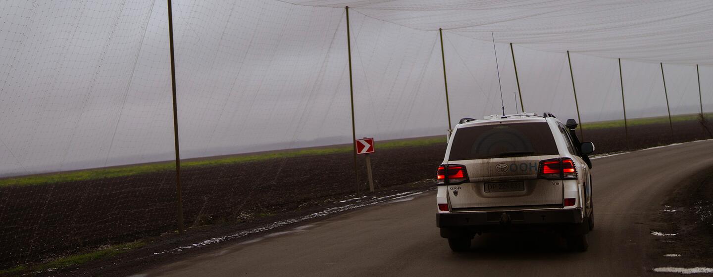Белый внедорожник едет по сельской дороге в Украине, проходя под большой сельскохозяйственной сетью, поддерживаемой полюсами.