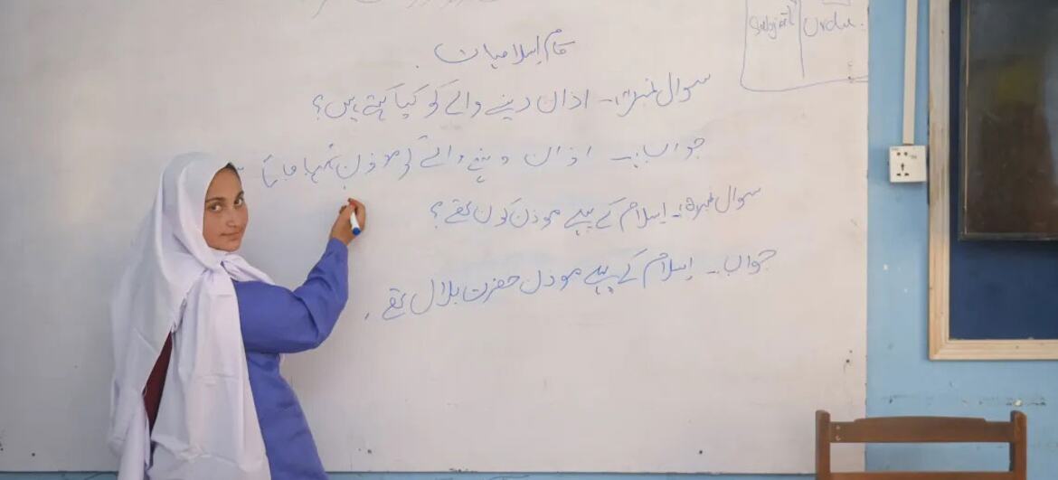 ایک سفید حجاب اور نیلے رنگ کی وردی میں ایک نوجوان لڑکی ایک کلاس روم میں وائٹ بورڈ کے سامنے کھڑی ہے، جو اردو میں لکھتی ہے۔ وائٹ بورڈ پر اردو متن ہے، اور دیوار پر یونیسیف اور ناروے کے نشان نظر آتے ہیں۔ یہ تصویر بلوچستان، پاکستان کی ایک دور دراز کمیونٹی میں لڑکیوں کے لئے سیکھنے اور بااختیار بنانے کا ایک لمحہ پیش کرتی ہے۔
