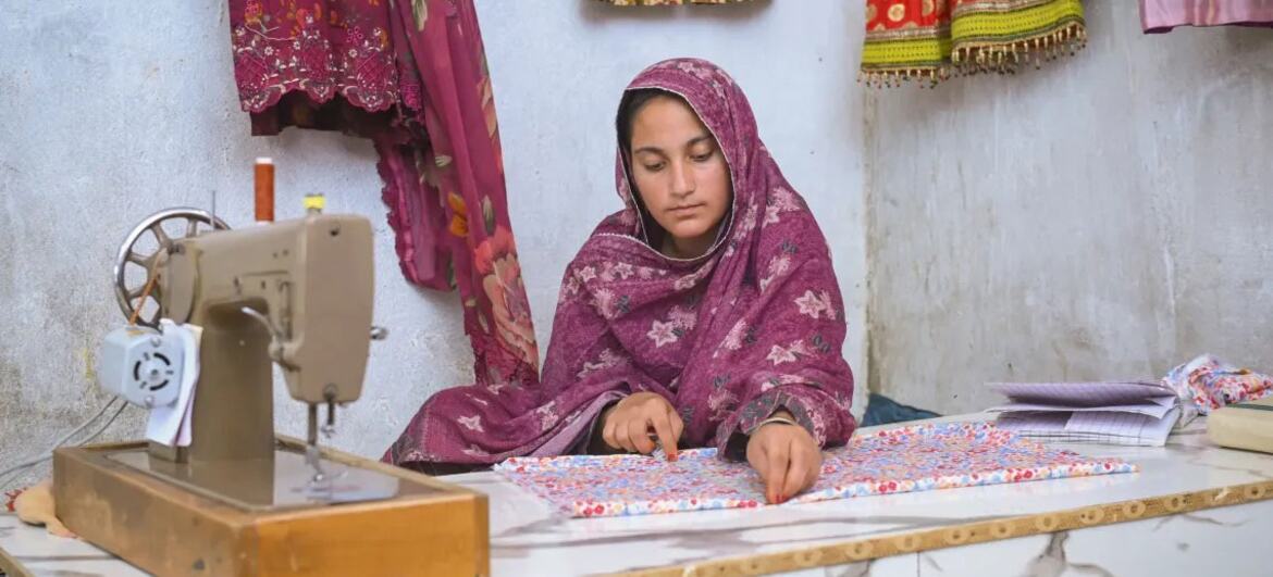 بلوچستان، پاکستان میں ایک معمولی گھر میں رنگین روایتی کپڑوں سے گھرا ہوا ایک نوجوان لڑکی جو پیلے رنگ کے ہیڈ شیلف پہنے ہوئی ہے وہ ایک پرانی سلائی مشین والی میز پر کپڑے سلائی کرنے پر توجہ مرکوز کر رہی ہے۔