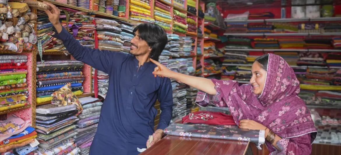 Uma menina com um lenço roxo aponta para um tecido numa loja de têxteis no Balochistão, no Paquistão, enquanto um homem faz gestos em direção a prateleiras cheias de têxteis coloridos.