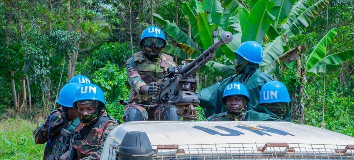 Tanzanian peacekeepers wearing blue UN helmets and camouflage uniforms are positioned on a military vehicle equipped with a mounted machine gun in a lush jungle setting.