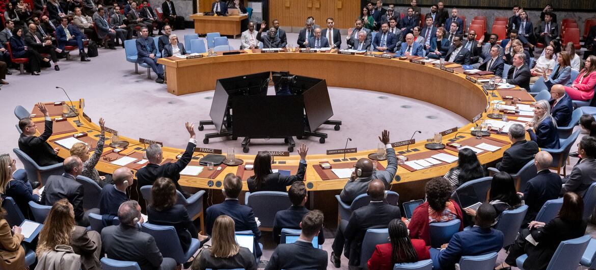 A wide-angle photograph of the United Nations Security Council chamber showing member states meeting to discuss the situation in the Middle East, with several delegates raising their hands.