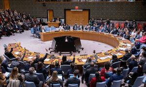 A wide-angle photograph of the United Nations Security Council chamber showing member states meeting to discuss the situation in the Middle East, with several delegates raising their hands.