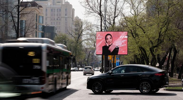 Большой розовый рекламный щит с портретом женщины и русским текстом стоит по правой стороне дороги, окруженный деревьями и зданиями.