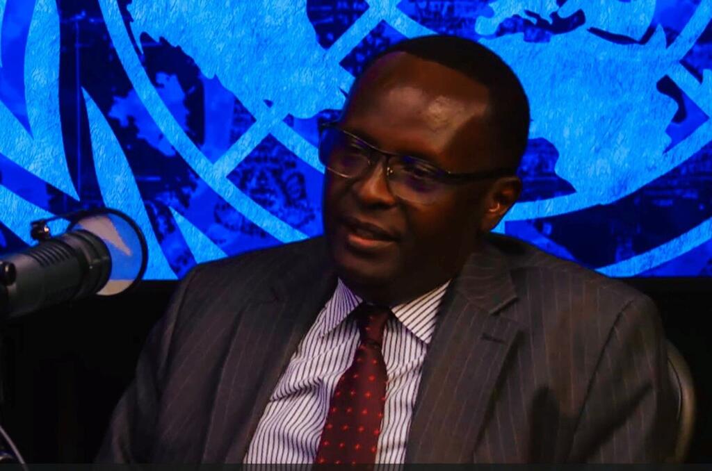 Mwanamume Mwafrika mwenye suti akizungumza kwenye kipaza sauti na ishara ya Umoja wa Mataifa yenye rangi ya bluu nyuma.