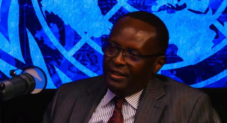 Mwanamume Mwafrika mwenye suti akizungumza kwenye kipaza sauti na ishara ya Umoja wa Mataifa yenye rangi ya bluu nyuma.