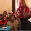 Mwalimu wa kike anayevaa hijab nyekundu amesimama katika darasa nchini Sudan, akifundisha kikundi cha wanafunzi wachanga walioketi kwenye meza zao.