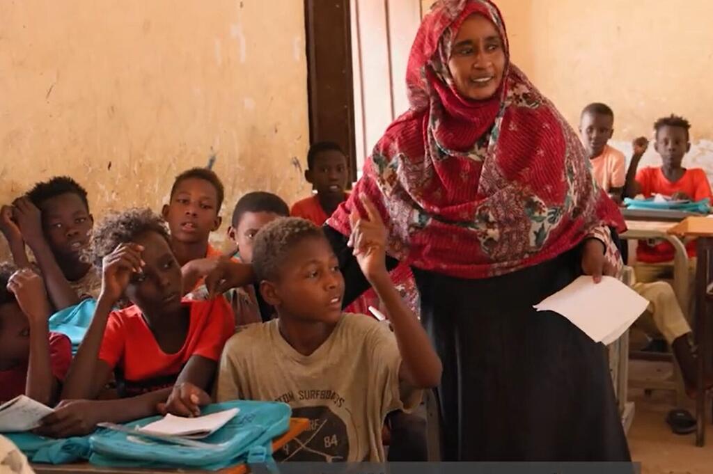 Mwalimu wa kike anayevaa hijab nyekundu amesimama katika darasa nchini Sudan, akifundisha kikundi cha wanafunzi wachanga walioketi kwenye meza zao.