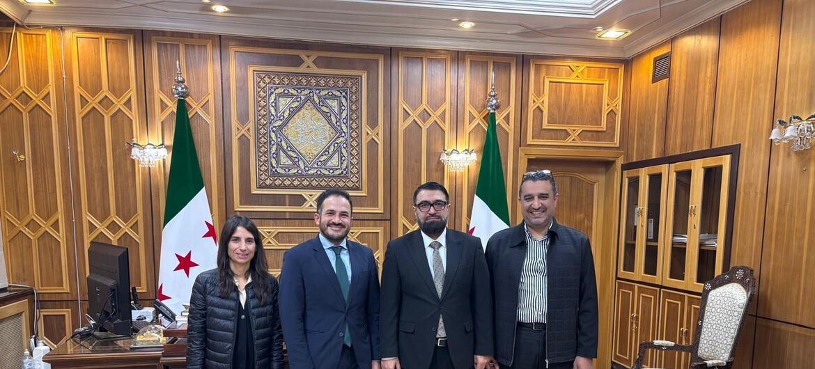 Four officials standing together in a wood-paneled room with Syrian flags in the background. From left to right: a female UN expert, two male UN experts, and the Syrian Minister of Justice.