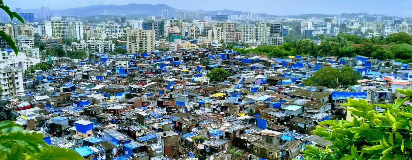 Aerial view of a densely packed slum area in Mumbai, India, with makeshift homes and blue tarps, contrasting with modern skyscrapers in the background.