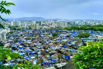 Vista aérea de una zona de barrios marginales densamente poblada en Mumbai, India, con viviendas improvisadas y lonas azules, en contraste con los rascacielos modernos del fondo.
