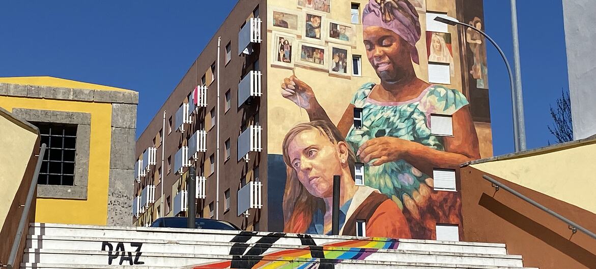 Um grande mural na lateral de um prédio de apartamentos em Zambujal, Portugal, retratando duas mulheres de diferentes etnias. Em primeiro plano, um conjunto de escadas brancas é pintado com um arco-íris e a palavra PAZ.