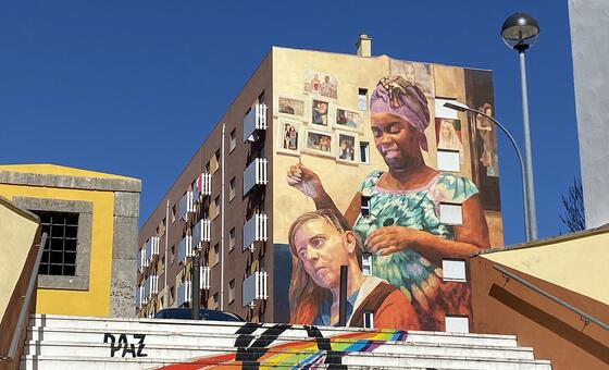 Um grande mural na lateral de um prédio de apartamentos em Zambujal, Portugal, retratando duas mulheres de diferentes etnias. Em primeiro plano, um conjunto de escadas brancas é pintado com um arco-íris e a palavra PAZ.
