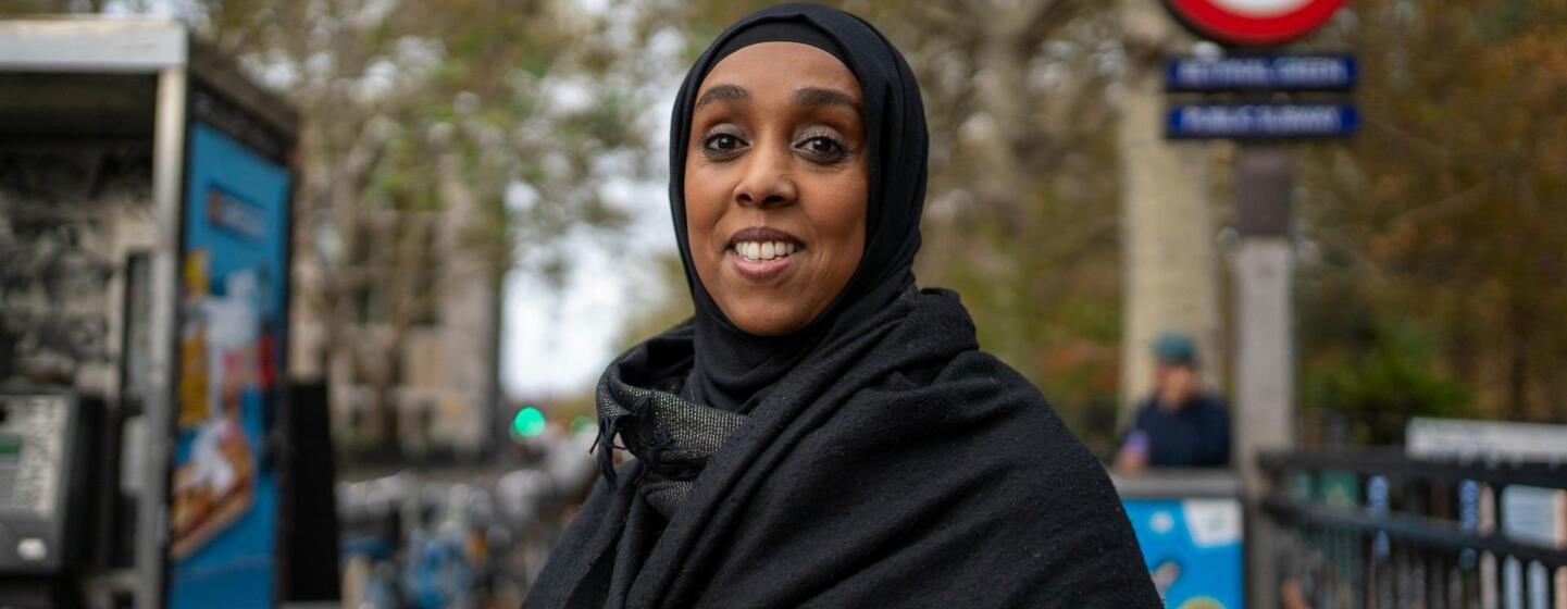 Safia Jama, a Somali-born activist in London, stands smiling in front of a Tube station, symbolizing her journey from Somalia to the UK and her commitment to community care.
