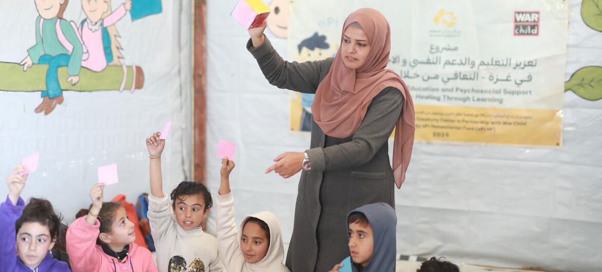 हिजाब पहने एक फिलिस्तीनी शिक्षक गाजा में एक अस्थायी सीखने की जगह में बच्चों की एक कक्षा को पढ़ाते हैं। बच्चे गुलाबी कार्ड के साथ एक शैक्षिक गतिविधि में अपना हाथ उठा रहे हैं, जिसे वॉर चाइल्ड द्वारा आयोजित किया गया है।