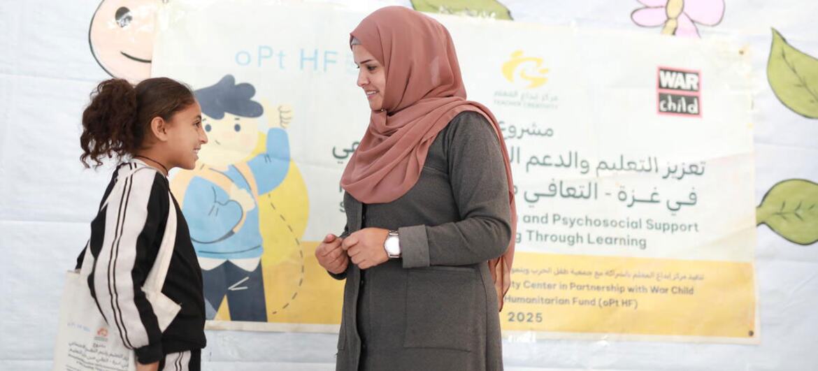 गाजा में एक अस्थायी सीखने की जगह में एक युवा लड़की के साथ बातचीत करते समय गुलाबी हिजाब में सल्वा नाम का एक फिलिस्तीनी शिक्षक मुस्कुराता है। उनके पीछे एक बैनर ओपीटी एचएफ और वॉर चाइल्ड के लोगो प्रदर्शित करता है।
