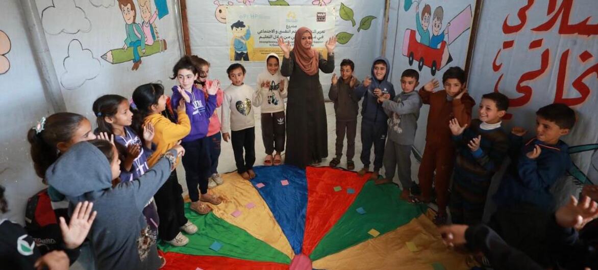 गाजा में एक अस्थायी सीखने की जगह के अंदर हिजाब पहनने वाली एक फिलिस्तीनी शिक्षिका बच्चों के एक समूह का नेतृत्व करती है। कमरे की दीवारों पर रंगीन चित्र हैं।