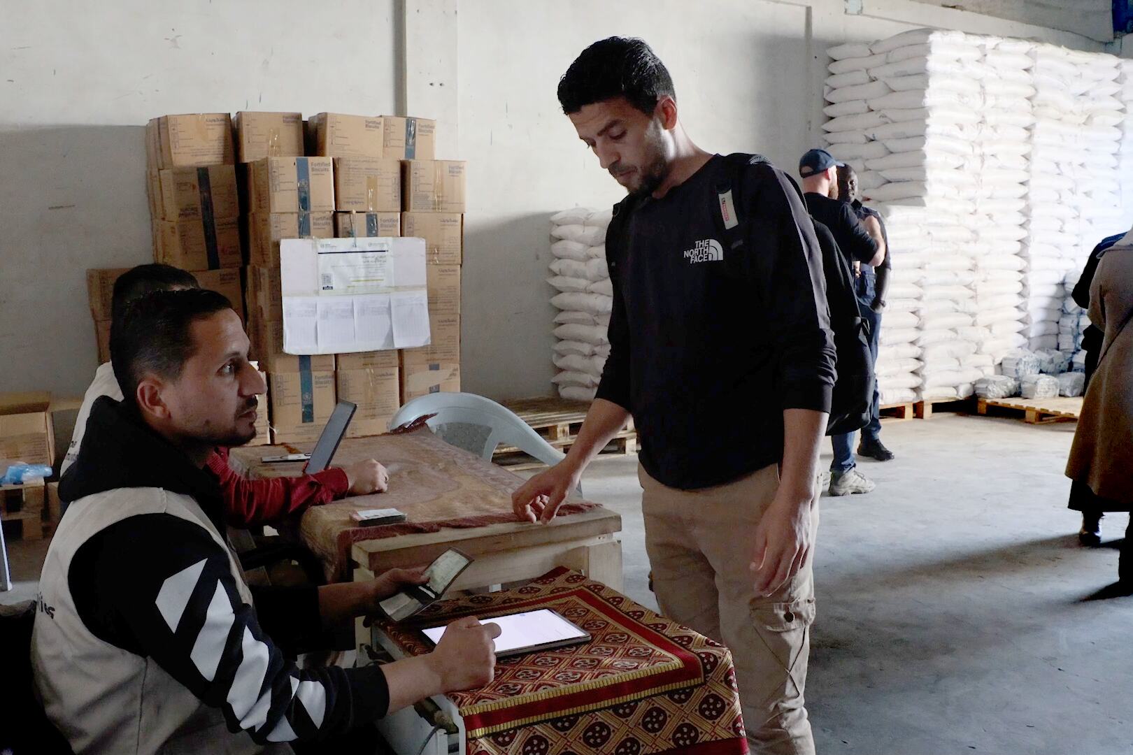 يقوم عامل مساعد في برنامج الأغذية العالمي بتسجيل مستلم في مركز توزيع الغذاء في مخيم الشاطئ للاجئين في غزة، باستخدام جهاز كمبيوتر لوحة. تتراكم أكوام من صناديق و أكياس المساعدات الغذائية في الخلفية.