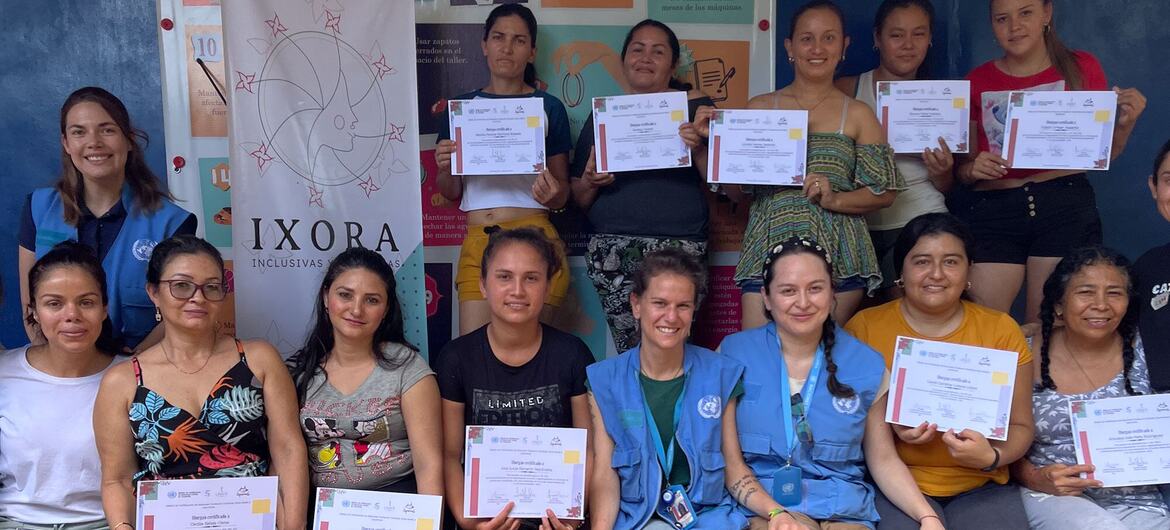 Un grupo de mujeres, entre ellas personal de la Misión de Verificación de la ONU en Colombia y diseñadoras de moda, posan juntas con certificados, sonrientes y de pie frente a un estandarte de IXORA Inclusive.