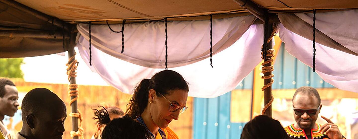 UN Under-Secretary-General Najla-Nassif Palma, wearing a yellow dress and glasses, interacts with a group of people under a tent at an outdoor event.