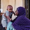 Uma mulher sorridente com um lenço roxo segura seu bebê menino em sua casa em Lomé, Togo.