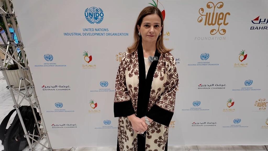 Mwanamke katika kanzu iliyo na muundo anasimama mbele ya mandhari inayoonyesha nembo za UNIDO, Bahrain Chamber, na WEIF2026 katika tukio huko Manama, Bahrain.
