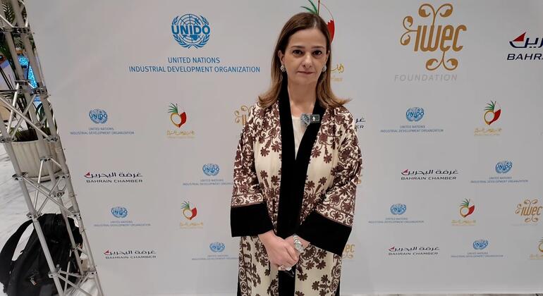 امرأة ترتدي معطفًا مرسومًا تقف أمام خلفية تضم شعارات UNIDO، غرفة البحرين، وWEIF2026 في حدث في ماناما، البحرين.
