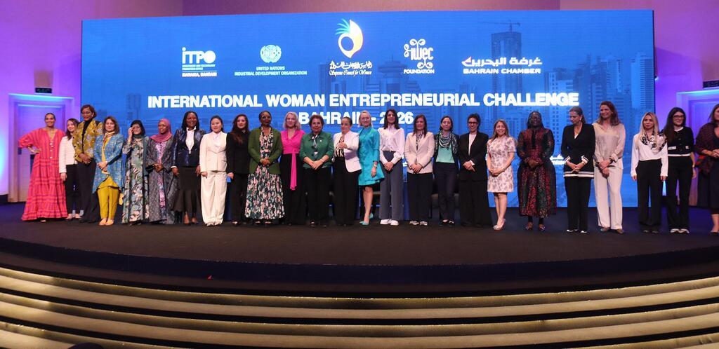 Kikundi cha wajasiriamali wa kike wanapiga picha kwenye jukwaa la International Woman Entrepreneurial Challenge 2026 huko Manama, Bahrain, na alama za tukio zimeonyeshwa kwenye skrini kubwa nyuma yao.