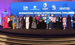 A group of women entrepreneurs pose for a photo on stage at the International Woman Entrepreneurial Challenge 2026 in Manama, Bahrain, with event logos displayed on a large screen behind them.