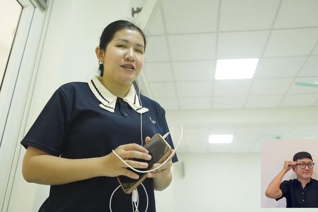 Mwanamke katika Navy bluu polo shati na kola nyeupe ana smartphone na nyeupe earphones plugged katika. Yeye ni kuzungumza, pengine wakati wa mahojiano au uwasilishaji. Katika background, mtu amevaa miwani ni kuonyeshwa katika picha ndogo inset, kurekebisha miwani yake.