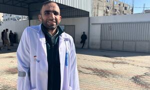 Daktari wa kiume mwenye tabasamu katika koti jeupe amesimama nje ya Hospitali ya Al-Shifa huko Gaza, pamoja na watu wengine na majengo yaliyoharibiwa yanayoonekana nyuma.