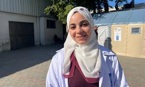 Mfanyakazi wa kike wa afya katika koti jeupe na hijab amesimama nje ya Hospitali ya Al-Shifa huko Gaza, na ishara za UNDP zinaonekana nyuma.
