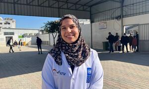 Mwanamke mwenye tabasamu ambaye ni mfanyakazi wa afya amevaa kanzu nyeupe iliyo na nembo za UNDP na KfW amesimama nje ya hospitali ya Al-Shifa huko Gaza. Watu wanaonekana nyuma.