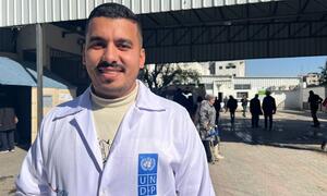 Mfanyakazi wa afya wa kiume mwenye tabasamu amevaa koti nyeupe la maabara na nembo ya UNDP amesimama nje ya Hospitali ya Al-Shifa huko Gaza. Watu wanaonekana nyuma.