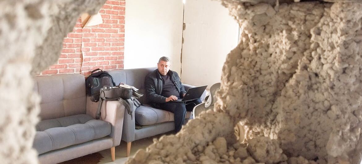 A man works on a laptop inside a damaged radio station in Gaza, viewed through a broken wall. This scene captures Zaman FM's return to broadcasting despite severe infrastructure damage and resource shortages.