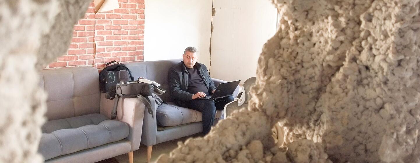 A man works on a laptop inside a damaged radio station in Gaza, viewed through a broken wall. This scene captures Zaman FM's return to broadcasting despite severe infrastructure damage and resource shortages.