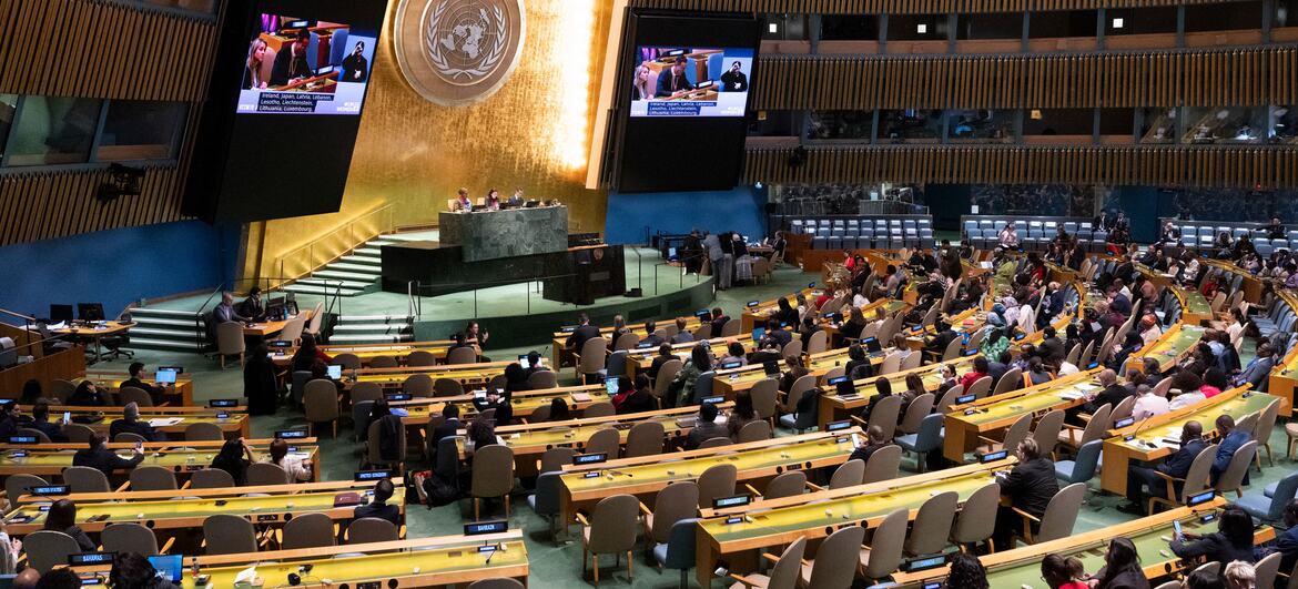 Uma vista panorâmica da sessão setenta da Comissão sobre o Estatuto da Mulher (CSW) na sede das Nações Unidas em Nova York. O grande salão de assembleias está cheio de delegados sentados em mesas curvas voltadas para um pódio central. Dois grandes ecrãs exibem os procedimentos. O emblema da ONU é exibido na parede atrás do pódio.