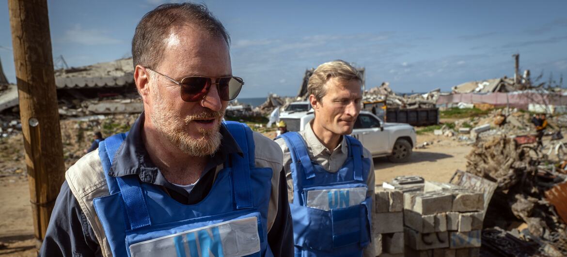 Shaun Hughes, WFP country director in Gaza, wearing a blue UN vest and sunglasses, walking through a site of destruction and rubble.