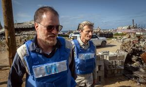Shaun Hughes, WFP country director in Gaza, wearing a blue UN vest and sunglasses, walking through a site of destruction and rubble.