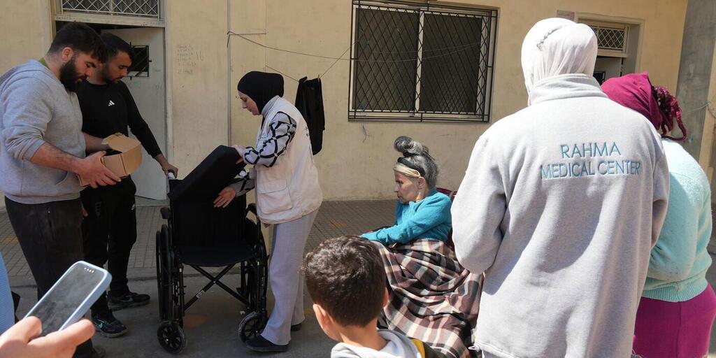 Los socios, con el apoyo del Fondo Humanitario para el Líbano administrado por OCHA, proporcionan ayudas de movilidad a las personas desplazadas en refugios del norte del Líbano el 10 de marzo de 2026. Una escena en el Centro Médico Rahma en el Líbano, donde se reúnen trabajadores humanitarios y miembros de la comunidad. Una mujer con un chaleco blanco asiste a una persona en silla de ruedas, mientras que una anciana está sentada envuelta en una manta. Una persona en primer plano lleva una sudadera gris con el nombre del centro en la espalda. Esta imagen se relaciona con la crisis humanitaria y la escalada militar en Oriente Medio.