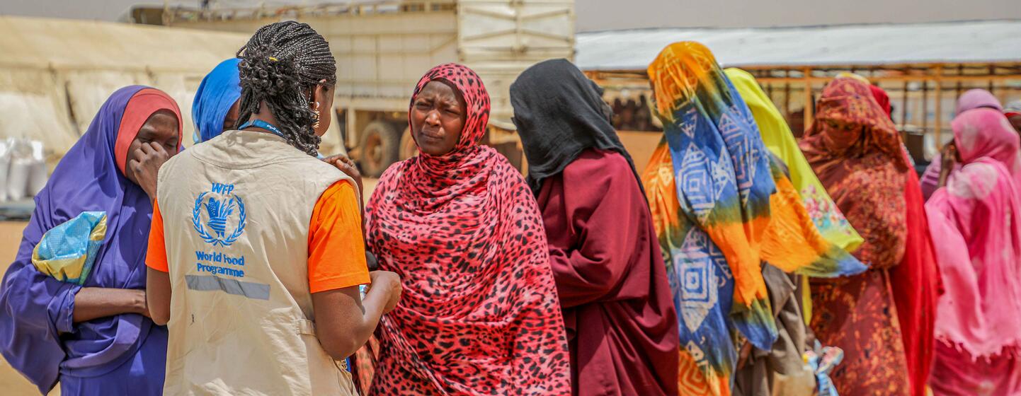Un travailleur humanitaire du Programme alimentaire mondial, vêtu d'un gilet beige arborant le logo du PAM, s'entretient avec une file de femmes portant des hijabs colorés à un point de distribution d'aide alimentaire à Adré, au Tchad. Les femmes attendent après un test biométrique visant à confirmer leur identité pour l'aide. Date : 18 mars 2026.