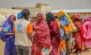 A World Food Programme aid worker in a beige vest with the WFP logo speaks with a line of women wearing colorful hijabs at a food aid distribution point in Adre, Chad. The women are waiting after a biometric test to confirm their identity for aid. Date: 18 March 2026.