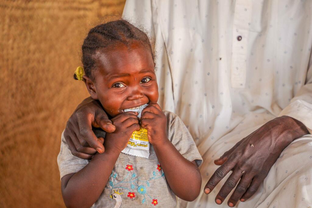 Un jeune enfant africain nommé Naba est montré en train de consommer des compléments alimentaires nutritionnels à Adré, Ouaddai, Tchad. Elle est tenue par les mains d'un adulte et sourit tout en mangeant le sachet de nourriture.