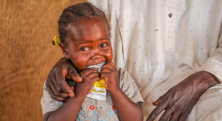 Маленькая африканская девочка по имени Наба ест питательные пищевые добавки в городе Адре, регион Уаддай, Чад. Её держат за руки взрослые, и она улыбается, потребляя содержимое пакета.