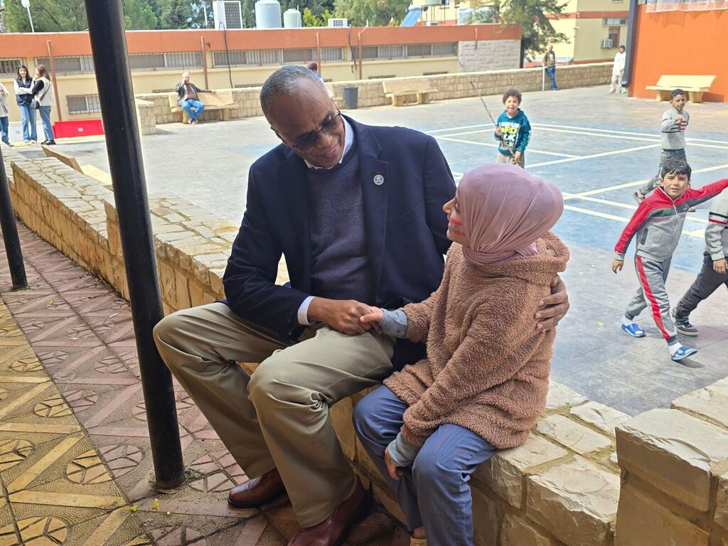Dr. Abdinasir Abubakar, Mwakilishi wa WHO nchini Lebanon, ameketi kwenye ukuta wa jiwe katika uwanja wa shule, akitabasamu na kushika mkono wa msichana mchanga aliyevaa hijab, huku watoto wengine wakicheza nyuma.