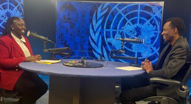 Mwanamke mweusi aliyevaa koti jekundu na mwanamume mweusi aliyevaa blazer nyeusi wameketi kwenye dawati katika studio, wakifanya mazungumzo kwa kutumia maikrofoni mbele ya picha kubwa ya alama ya Umoja wa Mataifa.